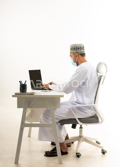 استخدام التقنيات الحديثه والعمل عن بعد، ارتداء الكمامة لاتخاذ الاجراءات الوقائية لفايروس كورونا, بورتريه من الجانب لرجل عربي خليجي عماني يرتدي الدشداشة والكمه العمانية التقليدية يجلس خلف طاولة خشبية يعمل على جهاز الحاسوب المحمول, خلفية بيضاء