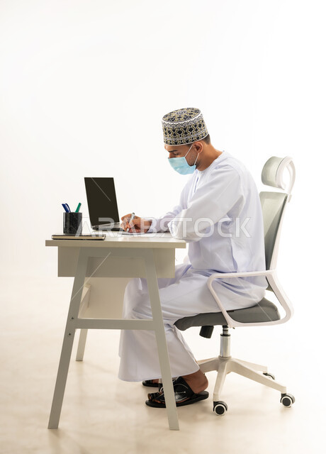 مفهوم الاجتهاد وانجاز الاعمال المطلوبة، ارتداء الكمامة لاتخاذ الاجراءات الوقائية لفايروس كورونا, بورتريه من الجانب لرجل عربي خليجي عماني يرتدي الدشداشة والكمه العمانية التقليدية يجلس خلف طاولة خشبية يقوم تدوين الملاحظات, خلفية بيضاء