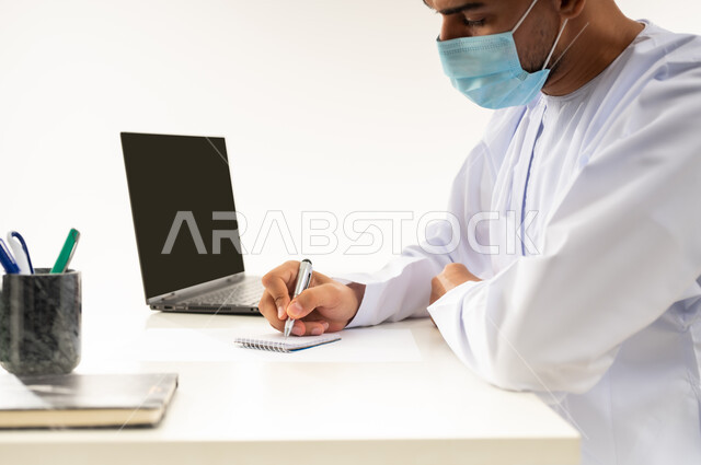 مفهوم الاجتهاد وانجاز الاعمال المطلوبة، ارتداء الكمامة لاتخاذ الاجراءات الوقائية لفايروس كورونا, بورتريه مقرب من الجانب لرجل عربي خليجي عماني يرتدي الدشداشة والكمه العمانية التقليدية يجلس خلف طاولة خشبية يقوم تدوين الملاحظات, خلفية بيضاء