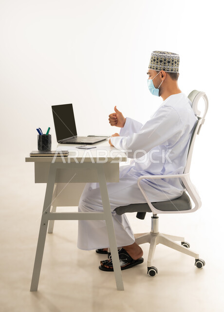 انجاز الاعمال المطلوبة بامتياز، ارتداء الكمامة لاتخاذ الاجراءات الوقائية لفايروس كورونا, بورتريه من الجانب لرجل عربي خليجي عماني يرتدي الدشداشة والكمه العمانية التقليدية يجلس خلف طاولة خشبية ينظر الحاسب المحمول بايماءات الاعجاب, خلفية بيضاء