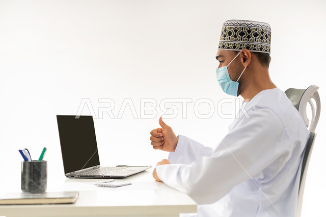 انجاز الاعمال المطلوبة بامتياز، ارتداء الكمامة لاتخاذ الاجراءات الوقائية لفايروس كورونا, بورتريه مقرب من الجانب لرجل عربي خليجي عماني يرتدي الدشداشة والكمه العمانية التقليدية يجلس خلف طاولة خشبية ينظر الحاسب المحمول بايماءات الاعجاب, خلفية بيضاء