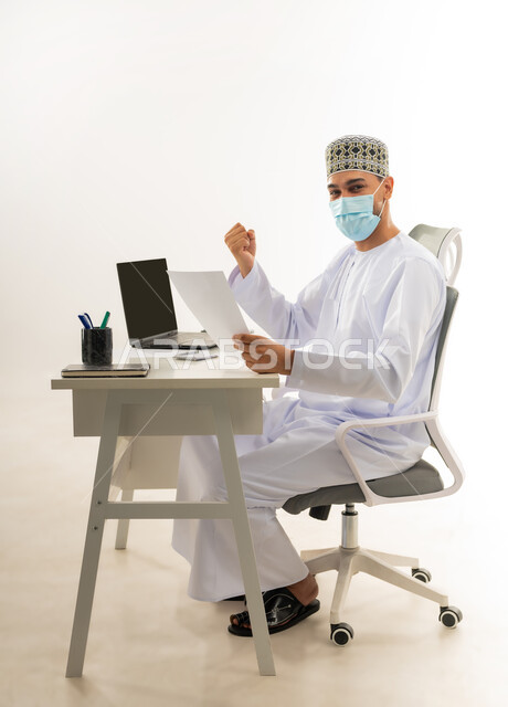 الفرح بالإنجاز وتحقيق الأهداف، ارتداء الكمامة لاتخاذ الاجراءات الوقائية لفايروس كورونا, بورتريه من الجانب لرجل عربي خليجي عماني يرتدي الدشداشة والكمه العمانية التقليدية يجلس خلف طاولة خشبية ينظر الى الكاميرا بايماءات النصر والفوز, خلفية بيضاء