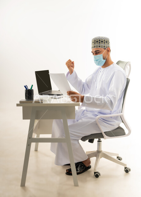 الفرح بالإنجاز وتحقيق الأهداف، ارتداء الكمامة لاتخاذ الاجراءات الوقائية لفايروس كورونا, بورتريه من الجانب لرجل عربي خليجي عماني يرتدي الدشداشة والكمه العمانية التقليدية يجلس خلف طاولة خشبية ينظر الى الورقة بايماءات النصر والفوز, خلفية بيضاء