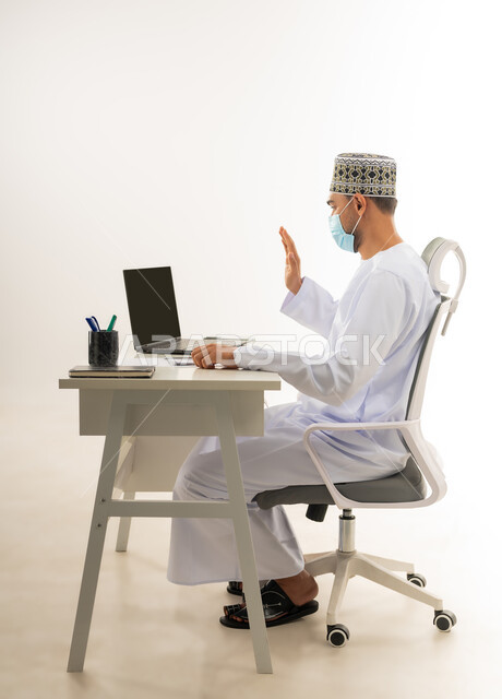 استخدام الاجهزة والتقنيات الحديثة, بورتريه لرجل عربي خليجي عماني يرتدي الدشداشة والكمة يجلس خلف طاولة يقوم باجراء مكالمة فيديو عبر جهاز الحاسب المحمول بايماءات القاء التحية والسلام، ارتداء الكمامة الطبية واتباع الاجراءات الوقائية ضد فايروس كورونا، خلفية بيضاء