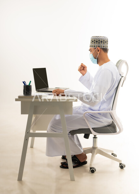 الفرح بالإنجاز وتحقيق الأهداف، ارتداء الكمامة لاتخاذ الاجراءات الوقائية لفايروس كورونا, بورتريه من الجانب لرجل عربي خليجي عماني يرتدي الدشداشة والكمه العمانية التقليدية يجلس خلف طاولة خشبية ينظر الى الحاسوب المحمول بايماءات النصر والفوز, خلفية بيضاء