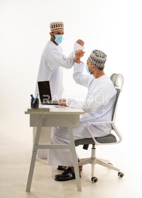 ارتداد الكمامة الطبية لاتخاذ الاجراءات الوقائية لفايروس كورونا, حسن الاستقبال بين زملاء العمل, اللطف والرقي في التعامل، بورتريه من الجانب لشابان عربيان خليجيان عمانيان يرتديان الدشداشة والكمه يقومان بالمصافحة بالاكواع، خلفية بيضاء