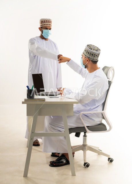ارتداد الكمامة الطبية لاتخاذ الاجراءات الوقائية لفايروس كورونا, حسن الاستقبال بين زملاء العمل, اللطف والرقي في التعامل، بورتريه من الجانب لشابان عربيان خليجيان عمانيان يرتديان الدشداشة والكمه يقومان بالمصافحة بقيضة اليد بايماءات السرور، خلفية بيضاء