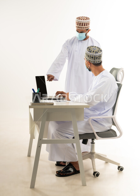 Wearing a medical mask to take preventive measures against the Corona virus, exchanging experiences and skills between employees, integrating modern technologies into practical life, portrait of an Arab Gulf Omani man wearing a dishdasha and a sleeve pointing his index finger to the laptop, white background