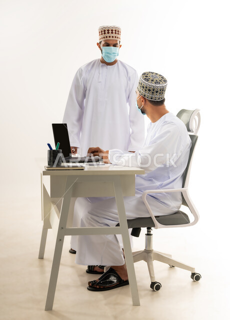 Participation in work among employees, wearing a medical mask to take preventive measures for the Corona virus, integrating modern technologies into practical life, side portrait of two Arab Gulf Omani men wearing a dishdasha and a kuma exchanging conversations and experiences, white background