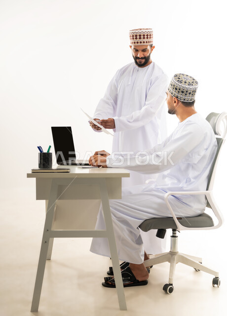 دمج التقنيات الحديثة بالحياة العملية, مهن ووظائف مكتبية, بورتريه لرجل عربي خليجي عماني يرتدي الدشداشة والكمه يقوم بإعطاء التعليمات وشرح المهام لزميله عبر الورق، مفهوم المشاركة في العمل، خلفية بيضاء