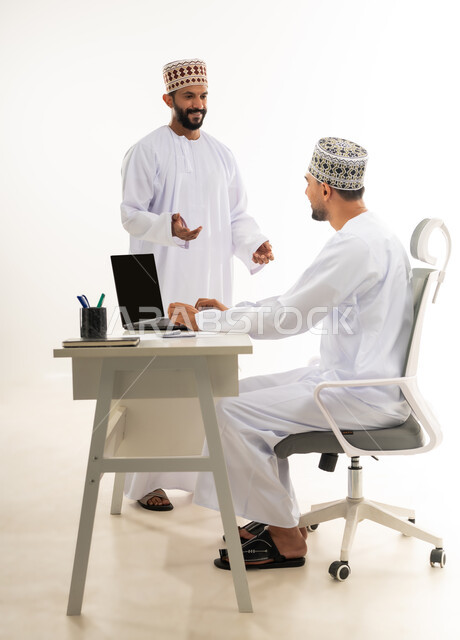 نقاشات وحوارات عمل بين الموظفين، دمج التقنيات الحديثة في الحياة العملية, بورتريه من الجانب لشابان عربيان خليجيان عمانيان يرتديان الدشداشة والكمه يقومان بتبادل الاحاديث بايماءات السعادة، خلفية بيضاء