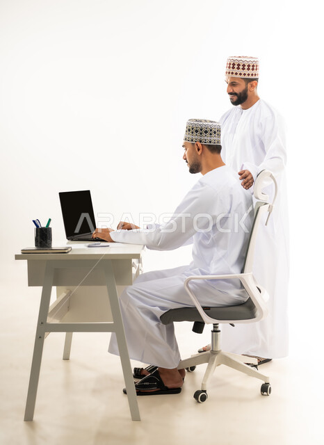 دمج التقنيات الحديثة في الحياة العملية, نقاشات وحوارات عمل بين الموظفين، بورتريه لشابان عربيان خليجيان عمانيان يرتديان الدشداشة والكمه ينظران الى جهاز الحاسوب الممحمول، خلفية بيضاء