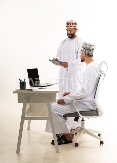 استخدام الاجهزة والتقنيات الحديثة, بورتريه من الجانب لرجل عربي خليجي عماني يرتدي الدشداشة والكمه يقوم بإعطاء التعليمات وشرح المهام لزميله، الرقي واللطف في التعامل بين الموظفين، مفهوم المشاركة في العمل، خلفية بيضاء