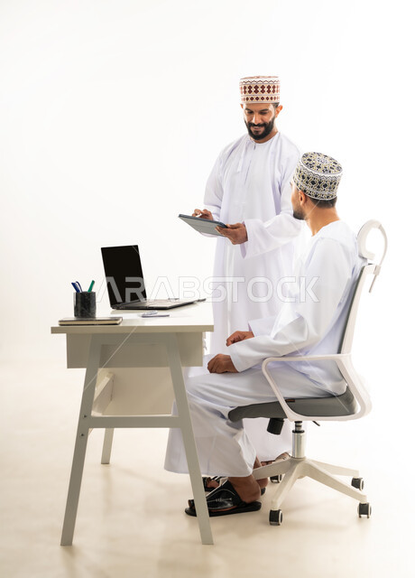 استخدام الاجهزة والتقنيات الحديثة, بورتريه من الجانب لرجل عربي خليجي عماني يرتدي الدشداشة والكمه يقوم بإعطاء التعليمات وشرح المهام لزميله، الرقي واللطف في التعامل بين الموظفين، مفهوم المشاركة في العمل، خلفية بيضاء