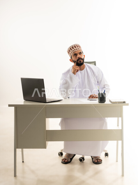 Looking up with gestures of thinking, distraction and being lost in thought about something, using modern and advanced technologies in work, portrait of a young Arab Gulf Omani man wearing a traditional dishdasha and kemeh sitting behind a wooden table working on a laptop, white background