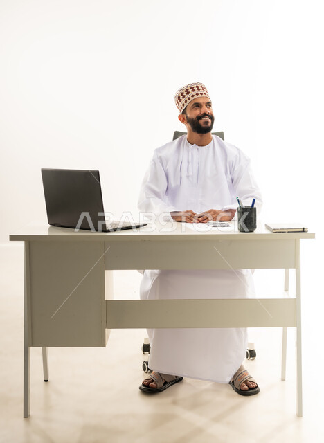 Using modern and advanced technologies in work, portrait of a young Arab Gulf Omani man wearing a traditional dishdasha and kummah sitting behind a wooden table working on a laptop, looking up with gestures of happiness, white background