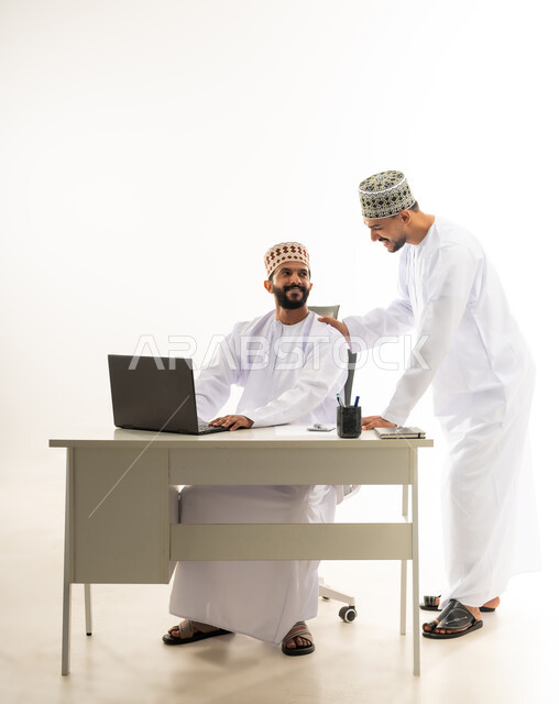 The concept of partnership and cooperation, exchange of experiences and skills, use of modern technical devices to accomplish tasks, portrait of two young Gulf Arab Omani men wearing the dishdasha and the kameh with expressions of joy and happiness, kindness and sophistication in dealing with employees, white background