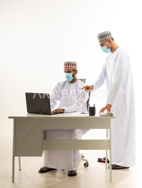 ارتداد الكمامة الطبية لاتخاذ الاجراءات الوقائية لفاروس كورونا, تبادل الخبرات و المهارات بين الموظفين, دمج التقنيات الحديثة في الحياة العملية، بورتريه لرجل عربي خليجي عماني يرتدي الدشداشة والكمه يشير باصبع السبابة الى الحاسب المحمول، خلفية بيضاء