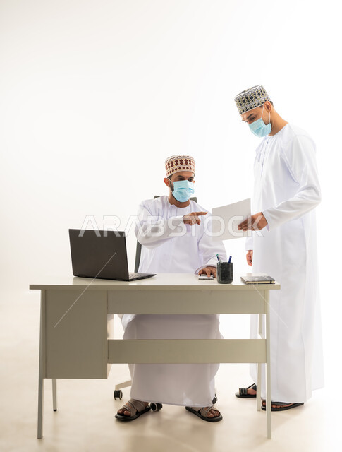 ارتداء الكمامة الطبية للحفاظ على الصحة, اتخاذ الاجراءات الوقائية لفايروس كورونا, بورتريه لرجل عربي خليجي عماني يرتدي الدشداشة والكمه يقوم بإعطاء التعليمات وشرح المهام لزميله، الرقي واللطف في التعامل بين الموظفين، مفهوم المشاركة في العمل، خلفية بيضاء