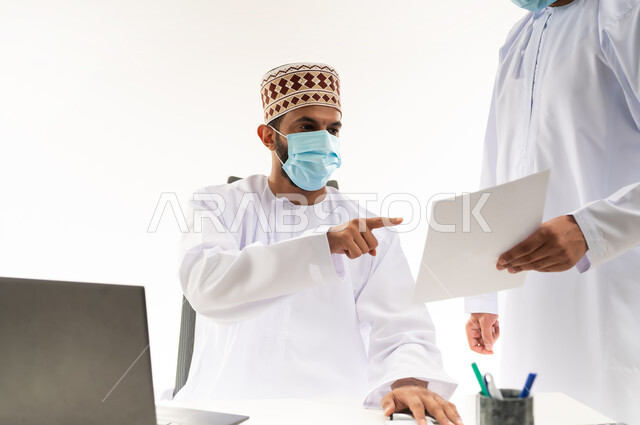 الرقي واللطف في التعامل بين الموظفين، ارتداء الكمامة الطبية للحفاظ على الصحة, اتخاذ الاجراءات الوقائية لفايروس كورونا, بورتريه مقرب لرجل عربي خليجي عماني يرتدي الدشداشة والكمه يقوم بإعطاء التعليمات وشرح المهام لزميله، مفهوم المشاركة في العمل، خلفية بيضاء