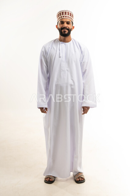 الاهتمام بالأناقة والمظهر الخارجي، التقاط الصور الشخصية للوثائق الرسمية، مفهوم الرجولة والثقة بالنفس، بورتريه مقرب لشاب عربي خليجي عماني مبتسم يرتدي الدشداشة و الكمه ينظر الى الكاميرا بإيماءات السرور والسعادة، صورة الجسم بالطول الكامل، خلفية بيضاء