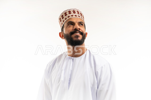 الاهتمام بالأناقة والمظهر الخارجي، التقاط الصور الشخصية للوثائق الرسمية، مفهوم الرجولة والثقة بالنفس، بورتريه مقرب لشاب عربي خليجي عماني مبتسم يرتدي الدشداشة و الكمه ينظر الى الاعلى بإيماءات السرور والسعادة، خلفية بيضاء