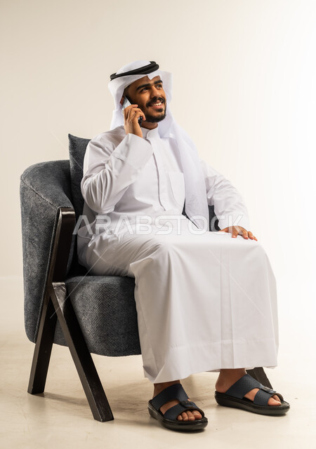 الرد على الاتصالات والمكالمات، التواصل مع الأصدقاء والاقارب، استخدام التقنيات الحديثة المتطورة، سماع أخبار سعيدة عبر الهاتف المحمول، بورتريه لشاب عربي خليجي كويتي يرتدي الثوب التقليدي والغتره يجلس على كرسي مريح يقوم بإجراء مكالمة هاتفية بايماءات السعاده، خلفيه بيضاء