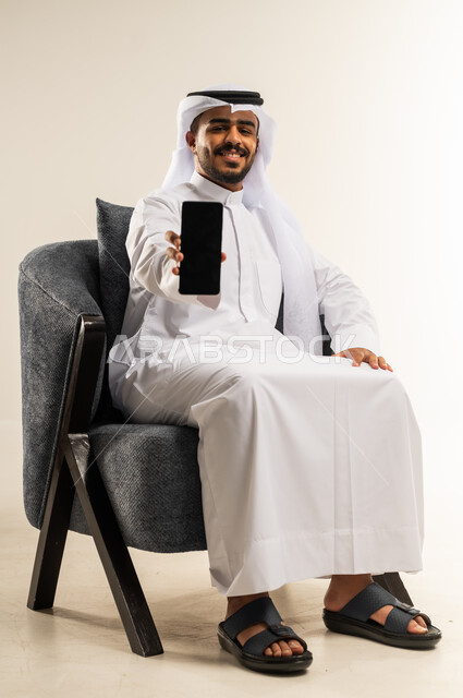 عرض تطبيقات وبرامج الجوال، بورتريه لرجل عربي خليجي كويتي يرتدي الثوب التقليدي والغتره يجلس على كرسي مريح يقوم بعرض شاشة سوداء فارغة عبر الهاتف المحمول، استخدام الاجهزة والتقنيات الحديثة المتطورة، النظر الى الكاميرا بتعابير السرور، خلفية بيضاء