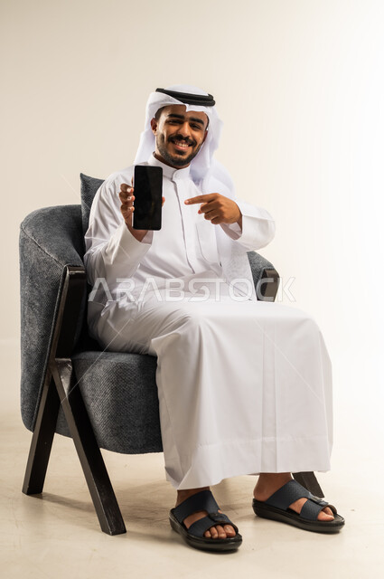 Using advanced modern devices and technologies, looking at the camera with expressions of pleasure, displaying mobile applications and programs, portrait of a Kuwaiti Gulf Arab man wearing a traditional dress sitting on a comfortable chair pointing his index finger at the empty black screen on a mobile phone, white background