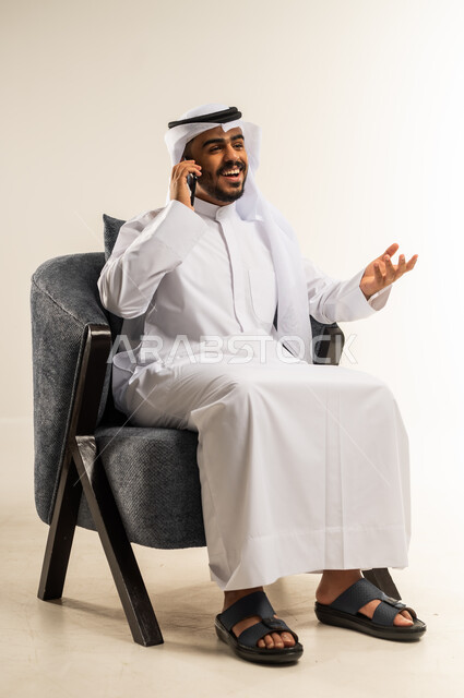 Communicating with friends and relatives, using advanced modern technologies, hearing happy news via mobile phone, portrait of a young Gulf Arab Kuwaiti man wearing traditional thobe sitting on a comfortable chair making a phone call and pointing upwards with gestures of happiness, white background