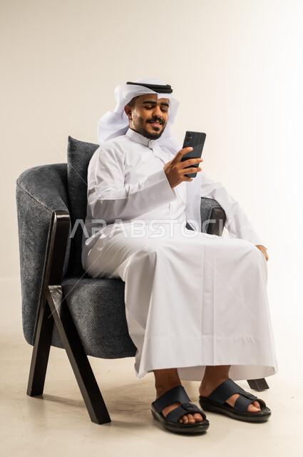 تصفح مواقع التواصل الاجتماعي, التواصل والدردشة مع الأهل والأصدقاء عبر تطبيقات الجوال, بورتريه لشاب عربي خليجي كويتي يرتدي الثوب التقليدي والغتره يجلس على كرسي مريح يقوم باستخدام الهاتف المحمول, استخدام التقنيات الحديثة المتطورة, خلفية بيضاء