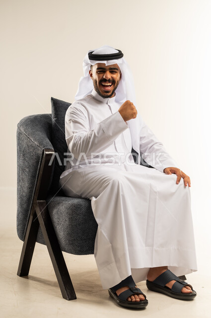 الانتصار والفوز والنجاح، مفهوم الانجاز وتحقيق الاهداف، بورتريه لشاب عربي خليجي كويتي يرتدي الثوب التقليدي والغتره يجلس على كرسي مريح ينظر الى الكاميرا يرفع قبضة يديه للاعلى بإيماءات الفرح والسرور, الشعور بالسعادة والقوة، خلفية بيضاء
