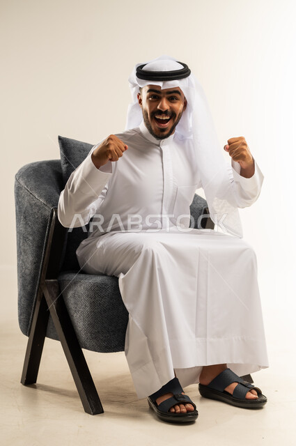 مفهوم الانجاز وتحقيق الاهداف، الانتصار والفوز والنجاح، بورتريه لشاب عربي خليجي كويتي يرتدي الثوب التقليدي والغتره يجلس على كرسي مريح ينظر الى الكاميرا يرفع قبضة يديه للاعلى بإيماءات الفرح والسرور, الشعور بالسعادة والقوة، خلفية بيضاء