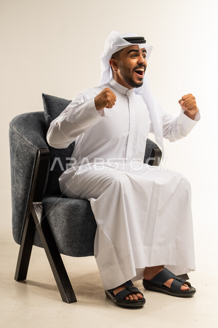 الشعور بالسعادة والقوة، مفهوم الانجاز وتحقيق الاهداف، الانتصار والفوز والنجاح، بورتريه لشاب عربي خليجي كويتي يرتدي الثوب التقليدي والغتره يجلس على كرسي مريح يرفع قبضة يديه للاعلى بإيماءات الفرح والسرور, خلفية بيضاء