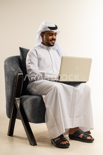إدارة شؤون الأعمال وتنظيمها، استخدام التقنيات الحديثة المتطورة، عقد إجتماع عمل عبر الإنترنت، بورتريه لشاب عربي خليجي كويتي يرتدي الثوب التقليدي والغتره يجلس على كرسي مريح يعمل عبر الحاسب المحمول, خلفية بيضاء