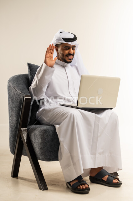 عقد إجتماع عمل عبر الإنترنت، مناقشة إستراتيجية المشروع, إدارة شؤون الأعمال وتنظيمها، استخدام التقنيات الحديثة المتطورة، بورتريه لشاب عربي خليجي كويتي يرتدي الثوب التقليدي يجلس والغتره على كرسي مريح يقوم بإجراء مكالمة فيديو من خلال الحاسب المحمول, خلفية بيضاء