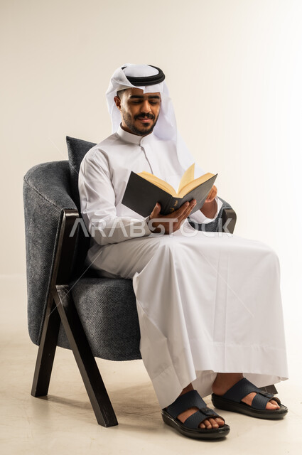 الاستمتاع بالأنشطة الترفيهية المفيدة في وقت الفراغ, مفهوم الثقافة والتعلم الذاتي, ممارسة هواية المطالعة, قراءة القصص والروايات, بورتريه لشاب عربي خليجي كويتي يرتدي الثوب التقليدي والغتره يجلس على كرسي مريح يمسك بيده كتاب بإيماءات السرور و السعادة, خلفية بيضاء