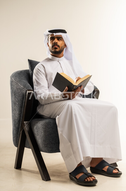 مفهوم الثقافة والتعلم الذاتي, ممارسة هواية المطالعة, الاستمتاع بالأنشطة الترفيهية المفيدة في وقت الفراغ, قراءة القصص والروايات, بورتريه لشاب عربي خليجي كويتي يرتدي الثوب التقليدي والغتره يجلس على كرسي مريح يحمل بيده كتاب ينظر الى شيء ما بإيماءات الجدية, خلفية بيضاء