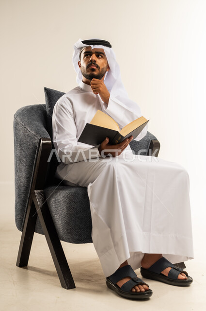 ممارسة هواية المطالعة, مفهوم الثقافة والتعلم الذاتي, الاستمتاع بالأنشطة الترفيهية المفيدة في وقت الفراغ, قراءة القصص والروايات, بورتريه لشاب عربي خليجي كويتي يرتدي الثوب التقليدي والغتره يجلس على كرسي مريح يحمل بيده كتاب يضع يده على ذقنه بإيماءات الحيرة, خلفية بيضاء