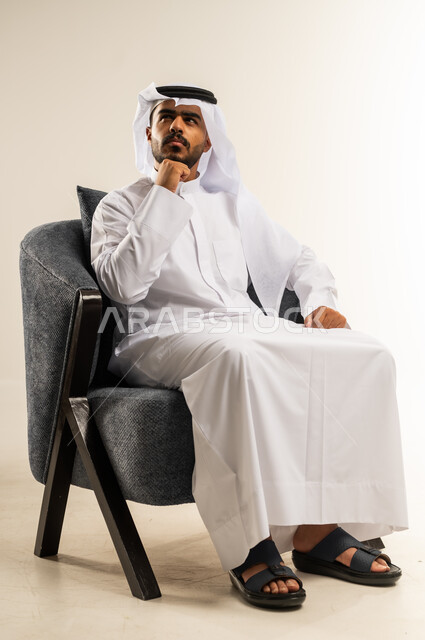 مفهوم شرود الذهن و التأمل والتخطيط للمستقبل, إيجاد حل لمشكلة ما, بورتريه لرجل عربي خليجي كويتي يرتدي الثوب التقليدي والغتره يجلس على كرسي مريح يضع يده على ذقنه بايماءات التفكير و الحيرة، خلفية بيضاء