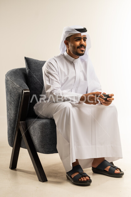 اجهزة وتقنيات الكترونية حديثة ترفيهيه, قضاء وقت ممتع ومسلي، التفاعل والاندماج مع ألعاب الفيديو والبلايستيشن، بورتريه لشاب عربي خليجي كويتي يرتدي الثوب التقليدي والغتره يجلس على كرسي مريح ويحمل بيده عصا التحكم، خلفية بيضاء