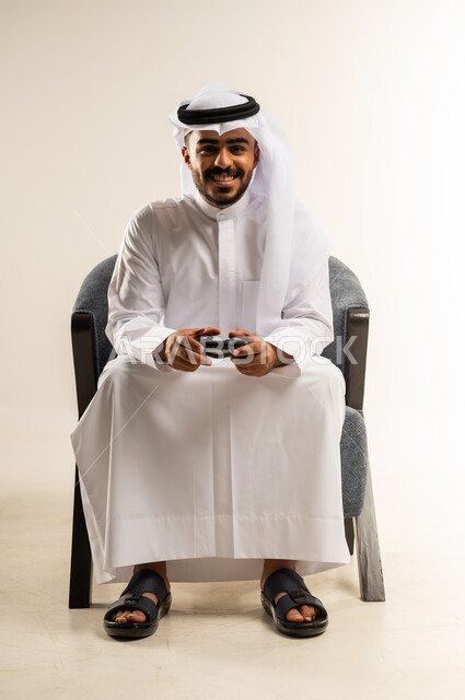 النظر الى الكاميرا بايماءات السرور والفرح, اجهزة وتقنيات الكترونية حديثة ترفيهيه, قضاء وقت ممتع ومسلي، التفاعل والاندماج مع ألعاب الفيديو والبلايستيشن، بورتريه لشاب عربي خليجي كويتي يرتدي الثوب التقليدي والغتره يجلس على كرسي مريح ويحمل بيده عصا التحكم، خلفية بيضاء