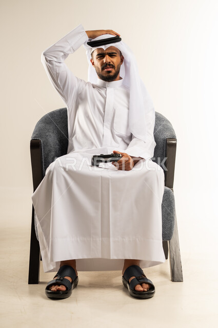 إيماءات الخسارة وخيبة الأمل، التسلية والترفيه بالفيديو جيمز في وقت الفراغ، استخدام الألعاب الالكترونية والأجهزة الحديثة، بورتريه لشاب عربي خليجي كويتي يرتدي الثوب التقليدي والغتره يمسك عصا التحكم ويضع يده على رأسه, النظر الى الكاميرا بنعابير الحسرة والحزن، خلفية بيضاء