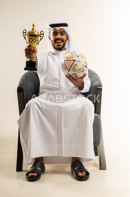 النظر الى الكاميرا بايماءات السعادة والفرح, مفهوم الربح والفوز، بورتريه لشاب عربي خليجي كويتي يرتدي الثوب التقليدي والغتره يجلس على كرسي مريح يحمل بيده كأس العالم الذهبي لرياضة كرة القدم، نشاط بدني للمحافظة على صحة الاجسام و اللياقة، خلفية بيضاء