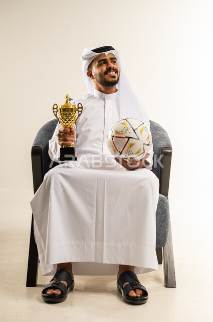 ايماءات السعادة والفرح, مفهوم الربح والفوز، بورتريه لشاب عربي خليجي كويتي يرتدي الثوب التقليدي والغتره يجلس على كرسي مريح يحمل بيده كأس العالم الذهبي لرياضة كرة القدم، نشاط بدني للمحافظة على صحة الاجسام و اللياقة، خلفية بيضاء