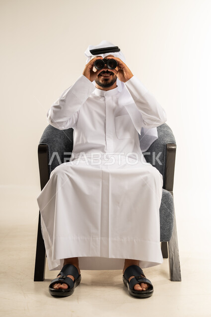 ايماءات الاستمتاع و السرور, رؤية الاشياء البعيدة و تقريبها, حب الاستكشاف و المغامرة, بورتريه لشاب عربي خليجي كويتي يرتدي الثوب التقليدي والغتره يجلس على كرسي مريح يحمل بيديه منظار ثنائي العينين و يينظر الى شيئ ما, خلفية بيضاء