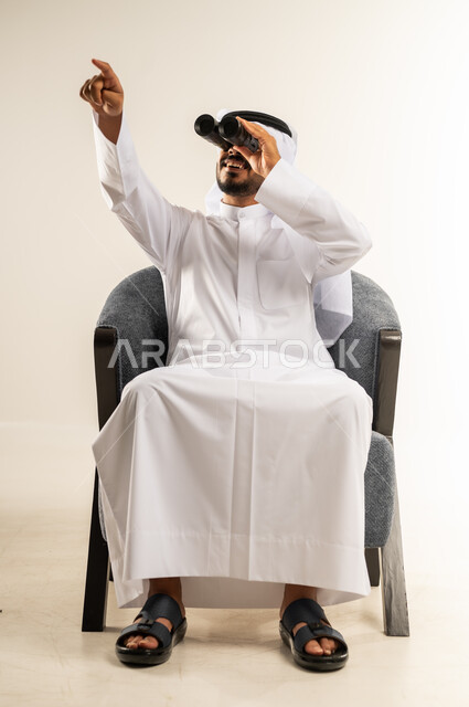 رؤية الاشياء البعيدة عبر المنظار، حب المغامرة والاستكشاف، بورتريه لشاب عربي خليجي كويتي يرتدي الثوب التقليدي والغتره يجلس على كرسي مريح ينظر من خلال المنظور ثنائي العينين يشير بإصبع السبابة الى شيء ما بإيماءات الفرح والسرور، خلفية بيضاء