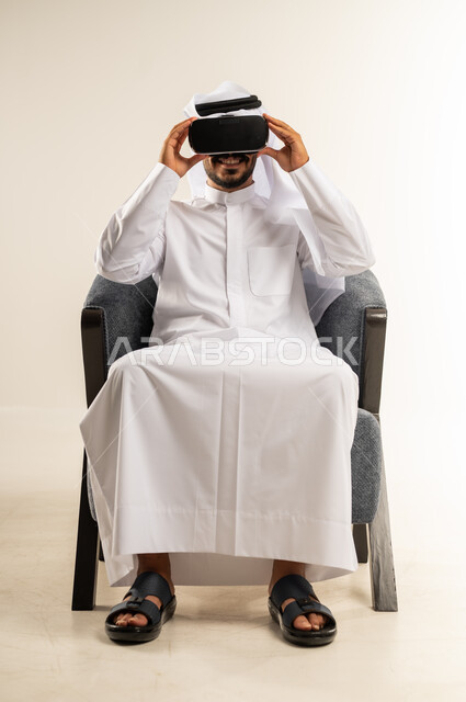 التطور التقني والتكنولوجي, المتعة و التسلية بالمشاهدة عبر نظارة الواقع الافتراضي، بورتريه لشاب عربي خليجي كويتي يرتدي الثوب التقليدي والغتره يجلس على كرسي مريح ينظر من خلال نظارات ثلاثية الأبعاد (الفي ار), الاندماج ومحاكاة العالم الخيالي، خلفية بيضاء