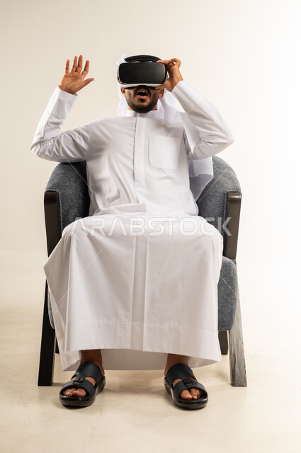 رفع كف اليد للأعلى بإيماءات الاندماج ومحاكاة العالم الخيالي، المتعة بالمشاهدة عبر نظارة الواقع الافتراضي، التطور التقني والتكنولوجي, بورتريه لشاب عربي خليجي كويتي يرتدي الثوب التقليدي والغتره يجلس على كرسي مريح ينظر من خلال نظارات ثلاثية الأبعاد (الفي ار), خلفية بيضاء