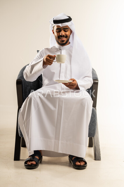 إيماءات السرور والسعادة, قضاء وقت الفراغ باحتساء المشروب المفضل, الجلوس والاستمتاع بالمشروبات الساخنة والأعشاب الشتوية الدافئة, بورتريه لشاب عربي خليجي كويتي يرتدي الثوب التقليدي والغتره يجلس على كرسي مريح يقوم باحتساء القهوة, خلفية بيضاء